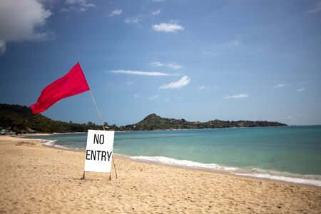 concept closed beach, private beach, not working. there is a red flag and a sign saying no entry on an empty tropical beach on a fine dayの写真素材
