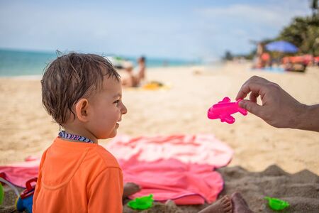 Concept Abolition Of Pandemic Quarantine. Start Of Flights Between Countries and travel. A little cute toddler sits on the beach, looks at a toy airplane held by a parent, and laughs happy.の写真素材