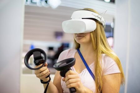 young caucasian girl in a virtual reality helmet holds control sticks in her hands and plays a game,...