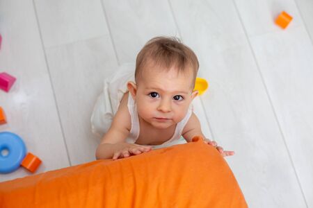little cute baby with brown eyes crawls on the orange corner of the bed, trying to stand on his feet and looking at the camera, top view, close-up. Against the background of a light floorの写真素材