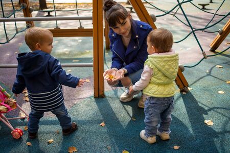 a young beautiful mother in a blue jacket squats in an autumn park in front of her little cute toddler son and daughter of 1 year old in blue clothes and smiles, showing him yellow leaves in his handsの写真素材