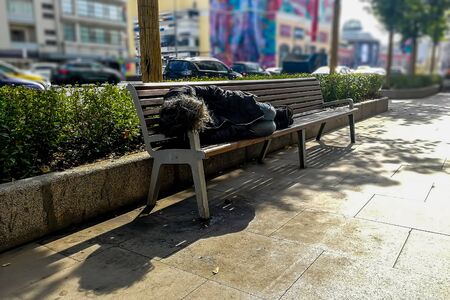 homeless beggar dirty man sleeping on a bench under a tree on the street, in the background the city, traffic, shopping center and colorful houses in blur. soft focus, close up.. の写真素材