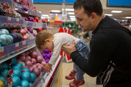 A man with love smile holds a small cute baby in his arms in a store of christmas new year decoration balls for christmas tree. baby is reaching down. close-up, soft focus, blur backgroundの写真素材