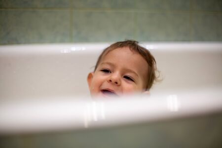 A cute Caucasian baby peeks out of the bathtub with smile, in the background is a green bathroom in...