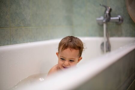 Cute Caucasian baby take a bath, playi in bath and smiles looking at water splashes, in the background a green bathroom in blur. close-up, soft focusの写真素材