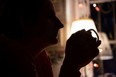 silhouette of a girl with closed eyes holding a white cup with a warm drink and drinking with pleasure. The concept of comfort and warmth. soft focus, dark window and christmas garland in blurの写真素材