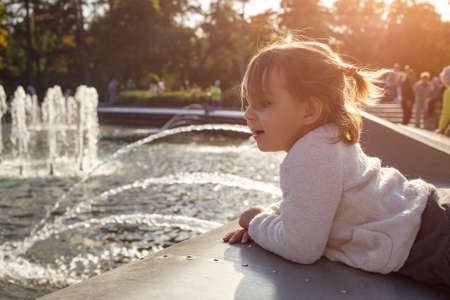 adorable toddler girl looks at the fountains in the park in the park on a sunny day.の写真素材