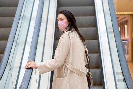 beautiful woman in a pink medical mask on her face against the background of the escadator in a public place.の写真素材