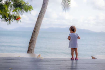 little toddler girl in a white dress looks at the sea while standing on the wooden floor outside. back view. soft focusの写真素材