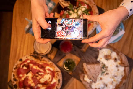 female hands takes pictures on a smartphone table with a delicious pizza in a restaurant. top viewの写真素材