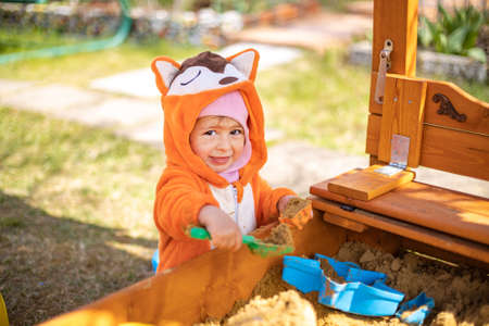 cute toddler in orange jumpsuit plays in the sand in the sandbox outdoors on a sunny dayの写真素材
