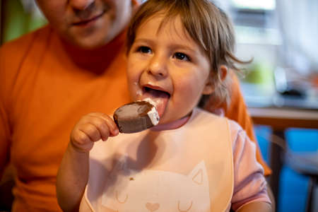 adorable toddler enjoys eating popsicle ice cream on parent's lap in home interior
