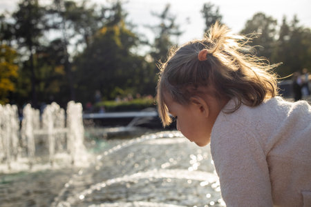 adorable toddler girl looks at the pond with fountains in the park on a sunny day. weekend family walk. spending time with children. artistic focusの写真素材