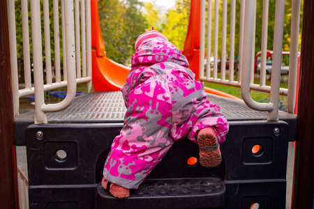 adorable kid in winter overalls climbs the steps of the children's slide on the playground. funny child climbs onto the slide on all fours rear viewの写真素材