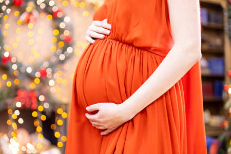 best christmas gift. faceless pregnant woman in a red dress hugs her belly against the background of the Christmas living room in blur. close-up no face.の写真素材