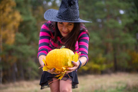 beautiful witch holds out a pumpkin to the camera in the autumn forestの写真素材