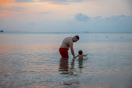 fat father with little child enter sea water rear view. people from behindの写真素材