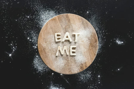 A round cookie with the words Eat Me written on it, surrounded by powder and dark background