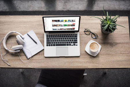A desk setup with a laptop, headphones, notebook, glasses, plant and coffee cup on a wooden table.