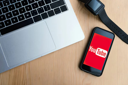 A laptop and smartphone displaying the YouTube app on a wooden desk setup.