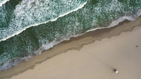 Bird's eye view of ocean waves crashing on a sandy beach with footprints