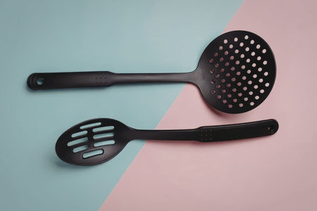Two black kitchen utensils, a slotted spoon and spatula, on pastel blue and pink background, clean and simple.