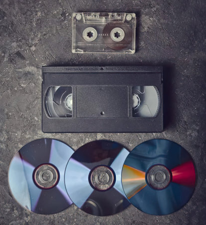 Vintage cassette tape and three compact discs on textured dark surface.