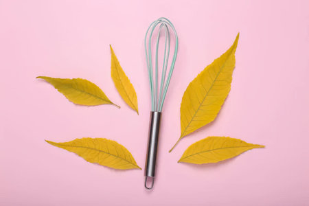 Metal whisk and yellow leaves on pink background, top view. Cooking utensils