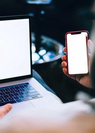 Mockup image of a woman's hand holding a smartphone with blank screen and a laptop on the background