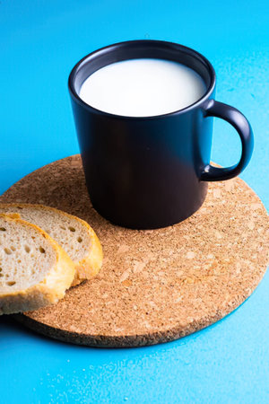 A black ceramic mug filled with milk placed on a cork coaster against a blue background.