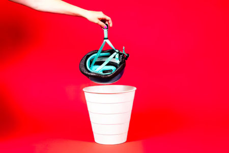 A person drops a blue bicycle helmet into a white trash can on a red background.