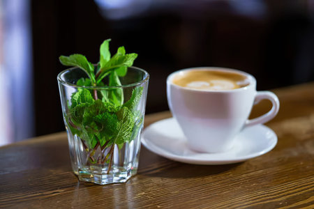 A clear glass with fresh green mint leaves and a cup of coffee on a wooden table.