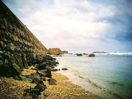 A peaceful coastal scene showing a rocky beach, green cliffs, and calm ocean waters under a partly cloudy sky.