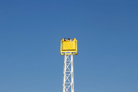 Minimalist photograph of visitors riding a tall amusement park attraction under a clear blue sky. Bright yellow observation cabin creates a strong visual contrast, symbolizing adventure, leisure, summer fun, freedom, and tourism.