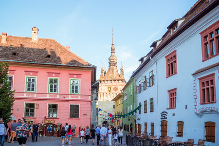 Sighisoara, Romania - August, 2019: Locals and tourists on the main square in medieval citadel of Sighisoara, restaurant and souvenir shops. Clock tower or tower of the Town Hall in the middle of cityのeditorial素材