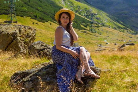 Smiling pretty girl in long dress sits on a rock holding a straw hat against the backgdrop of high mountains. Woman sitting near Transfegerasan, one of the most spectacular mountain road in the world.の写真素材