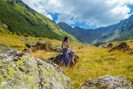 side photo of girl in long blue white dress sits on a rock and dreaming against the high mountains backgdrop. Woman sitting near Transfegerasan, one of the most spectacular mountain road in the worldの写真素材