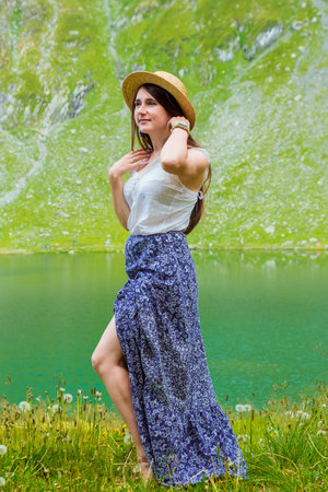 A beautiful woman in a long dress and straw hat stands on the shores of Lake Balea against the backdrop of the high Romanian mountains. exciting holidays in the mountains in Transylvania, Romaniaの写真素材