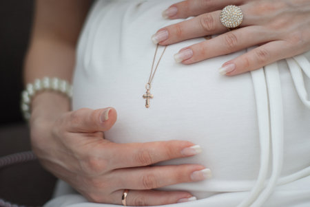 close up of pregnant woman in white dress touching her belly with hands and holding golden cross on the dark background. Happy pregnancy conceptの写真素材