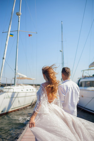 The newlyweds walk near the yacht club. The husbend leads the wife by the hand on the pier between the yachts. Wind waving the dress and the hair of the bride. Young couple back viewの写真素材
