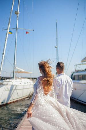 The newlyweds walk near the yacht club. The husbend leads the wife by the hand on the pier between the yachts. Wind waving the dress and the hair of the bride. Young couple back view