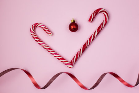 Christmas candies with ribbon on the pink background. Heart made from candy cane and christmas ball with wave of red ribbon on the pink paper. Merry Christmas. Happy Valentines Dayの写真素材