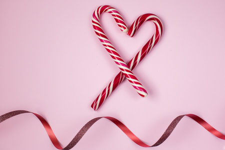 Christmas candies with ribbon on the pink background. Heart made from candy cane with wave of red ribbon on the pink paper. Merry Christmas. Happy Valentines Dayの写真素材
