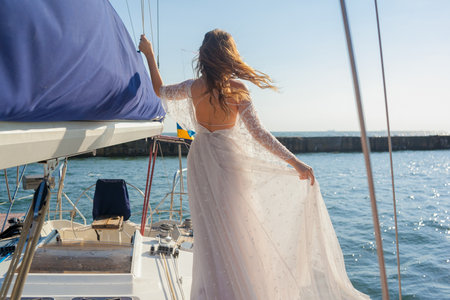 Bride holds the dress on the deck of the yacht. Wind waving the dress and the hair of the bride. Young woman back view. Wedding trip on a boatの写真素材