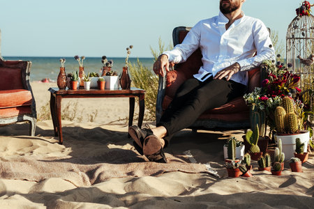 Cropped photo of the groom sitting on the chair on the wedding ceremony on the beach. relaxed man in a cozy setting for a date. wedding decoration with cactus and succulents on the seaside.の写真素材