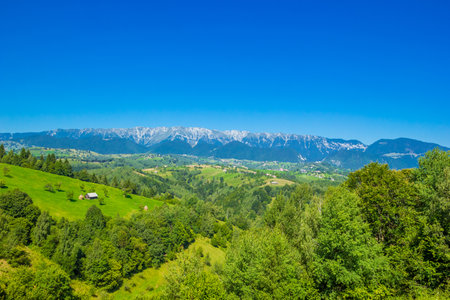 Stunning alpine landscape with green fields and high snowy Piatra Craiului mountains near Brasov. Mountain farm with old wooden house. Bran, Transylvania, Romania, Europe.の写真素材