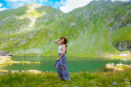 A beautiful woman in a long dress and straw hat stands on the shores of Lake Balea against the backdrop of the high Romanian mountains. exciting holidays in the mountains in Transylvania, Romaniaの写真素材