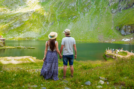 A beautiful woman in a long dress and straw hat stands with man on the shores of Lake Balea against the backdrop of the high Romanian mountains. exciting holidays in mountains in Transylvania, Romaniaの写真素材