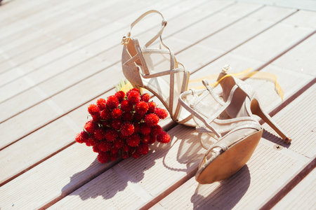 Bright shoes for stylish bride. high hill pair of stylish shoes and brides bouquet with red flowers on the wood backgroundの写真素材