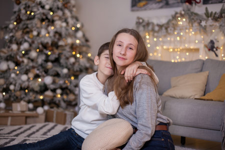 happy brother and sister sitting on floor near sofa and christmas tree with presents and looking at camera. Boy and girl at Christmas time. Merry Christmas and Happy New Yearの写真素材